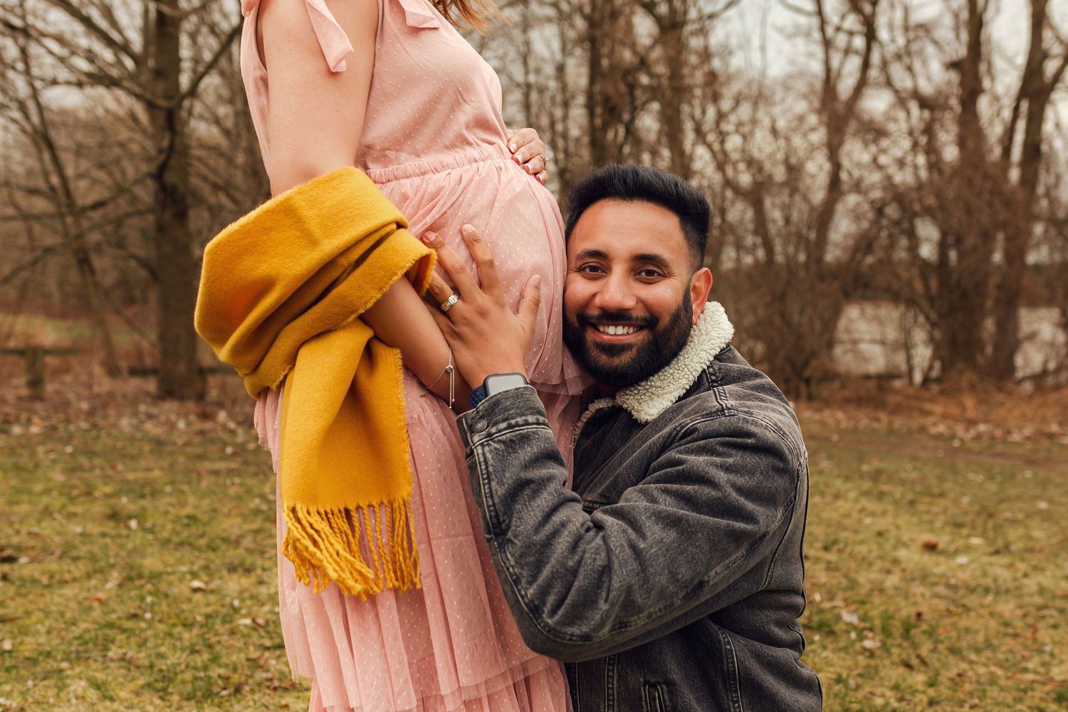 A joyful outdoor portrait showing someone in a denim jacket listening to the pregnant belly of a woman in a pink dress at Roth Park.
