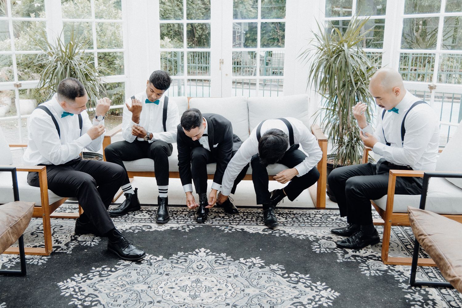 Groomsmen in black suits and suspenders getting ready in a bright sunroom captured by Apertured Media Auckland Wedding Photographer.