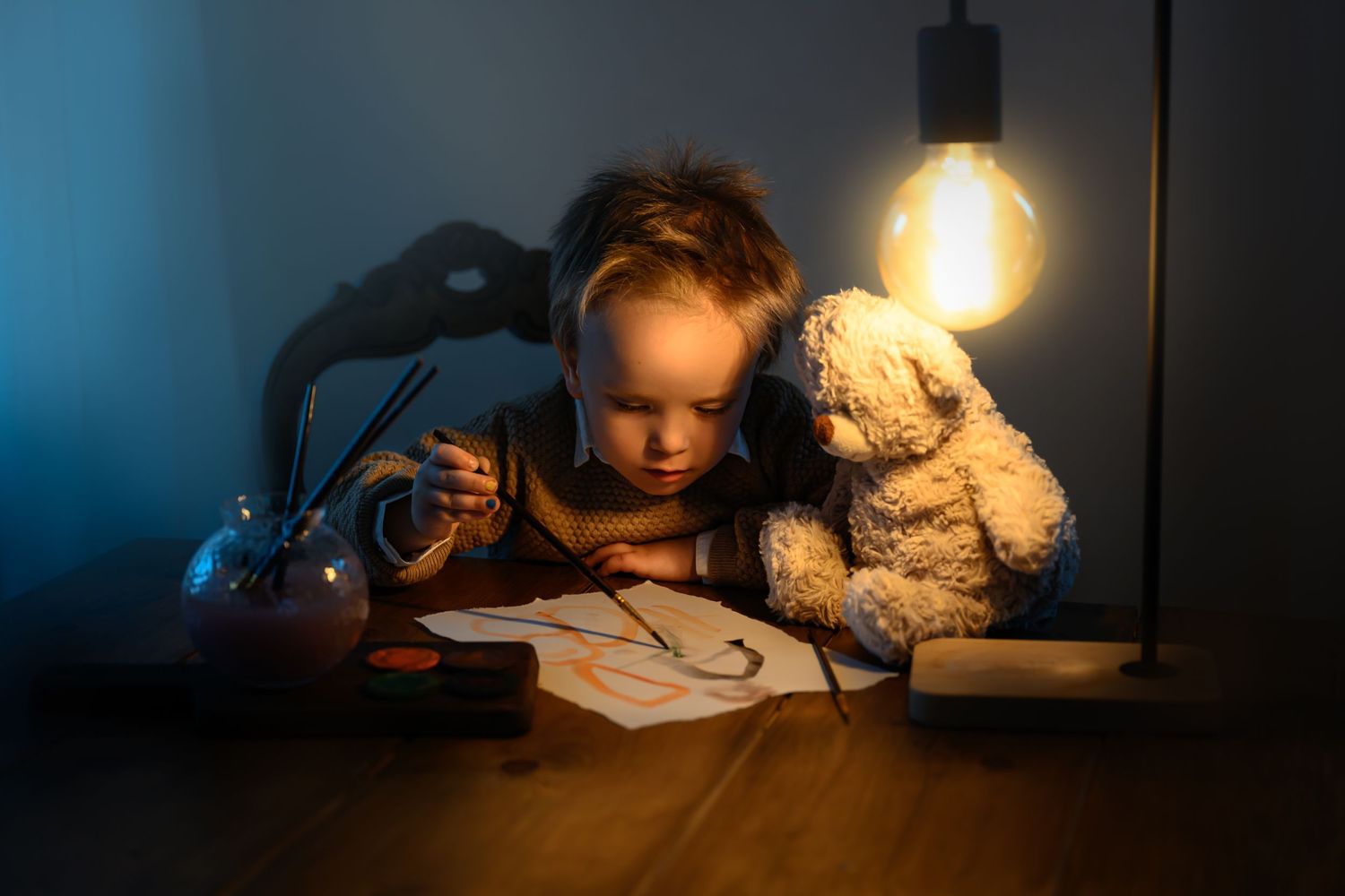 Vackert foto av en pojke som målar en teckning. Bredvid honom en nalle och en lampa.