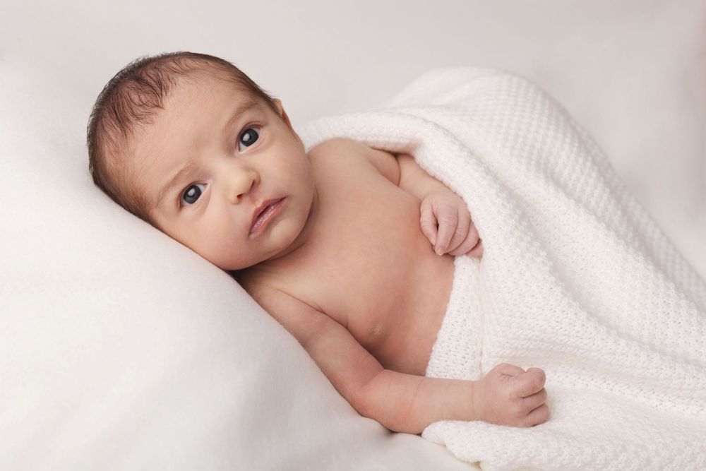 Sweet baby swaddled in white blanket on soft pillow looking at camera with bright eyes.