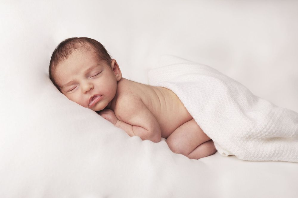 A sleeping newborn baby partially covered with white blanket rests peacefully on soft white bedding.