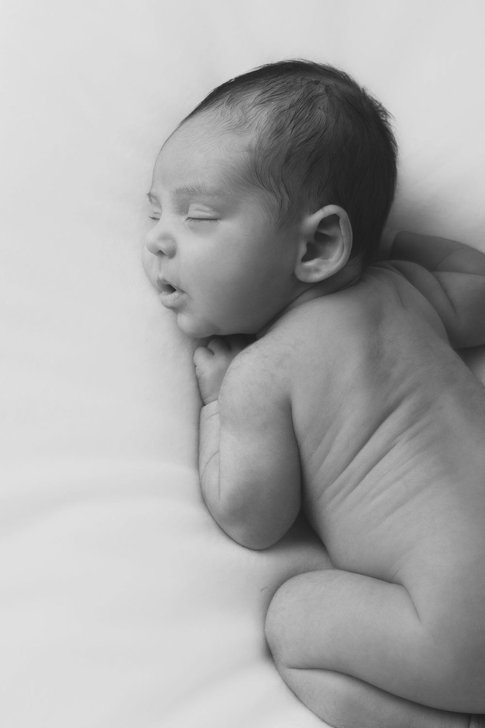 Black and white photo of a peaceful sleeping newborn baby lying on their side against a white background.
