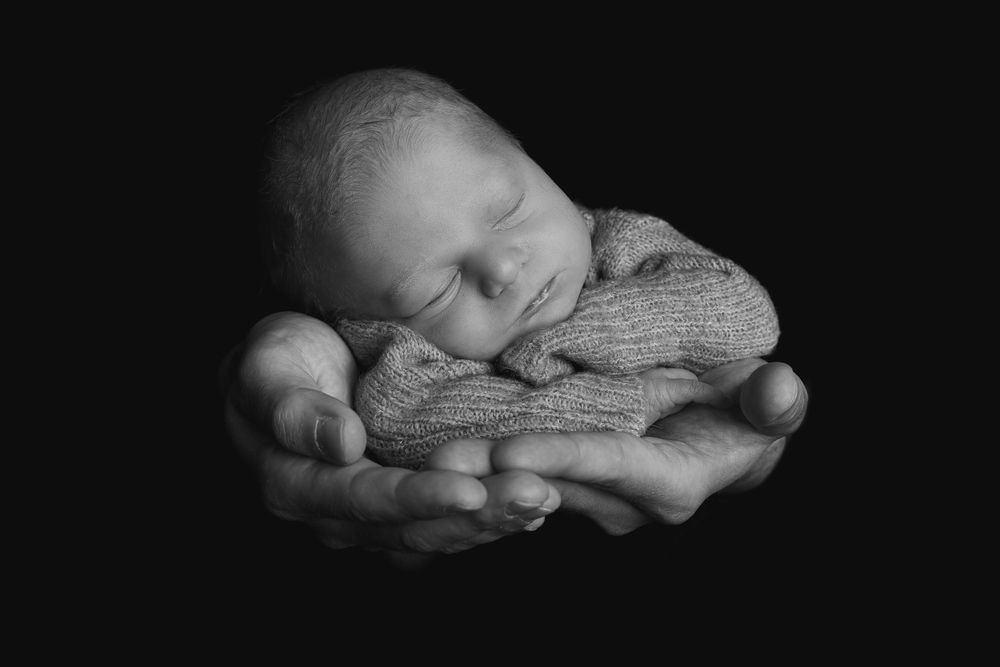 A sleeping newborn baby wrapped in knit fabric rests peacefully in cupped hands against a black background.