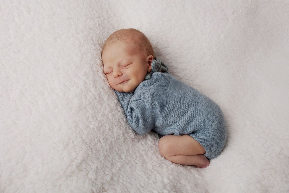 Sleeping newborn baby wearing cozy gray sweater curled up on soft white blanket during professional photography session.