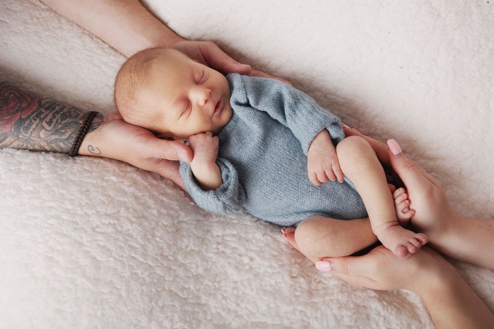 A sleeping newborn wearing a soft blue onesie lies gently cradled in caring hands on white textured fabric.