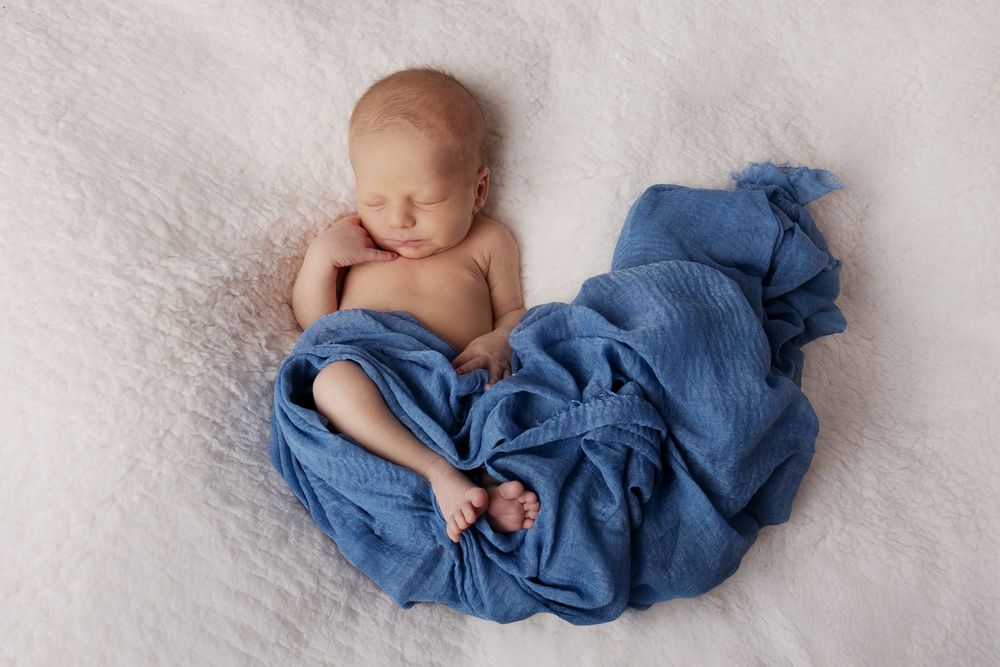 A sleeping newborn baby wrapped in soft blue swaddle fabric lying on white textured backdrop during professional photoshoot.