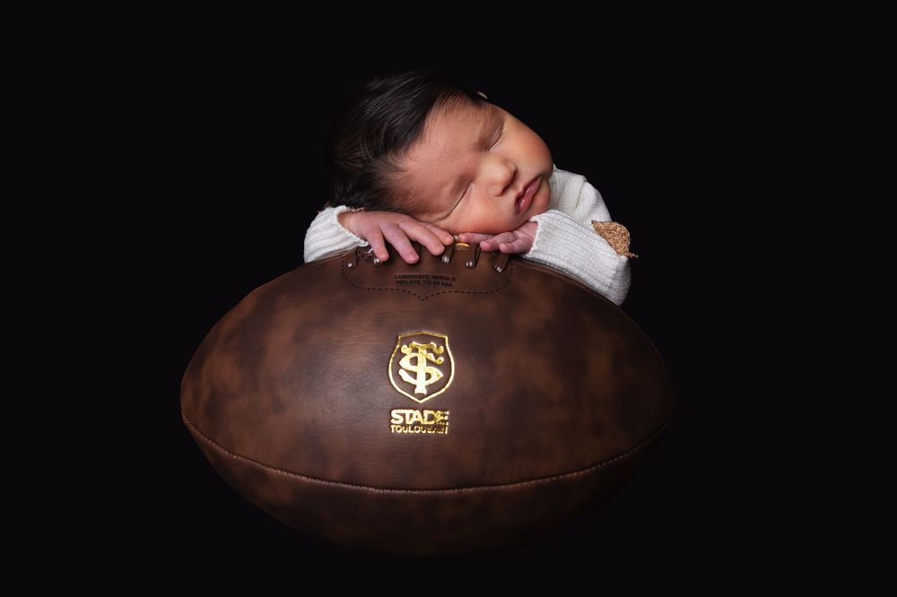 A sleeping newborn rests peacefully on a brown leather football with a gold emblem.