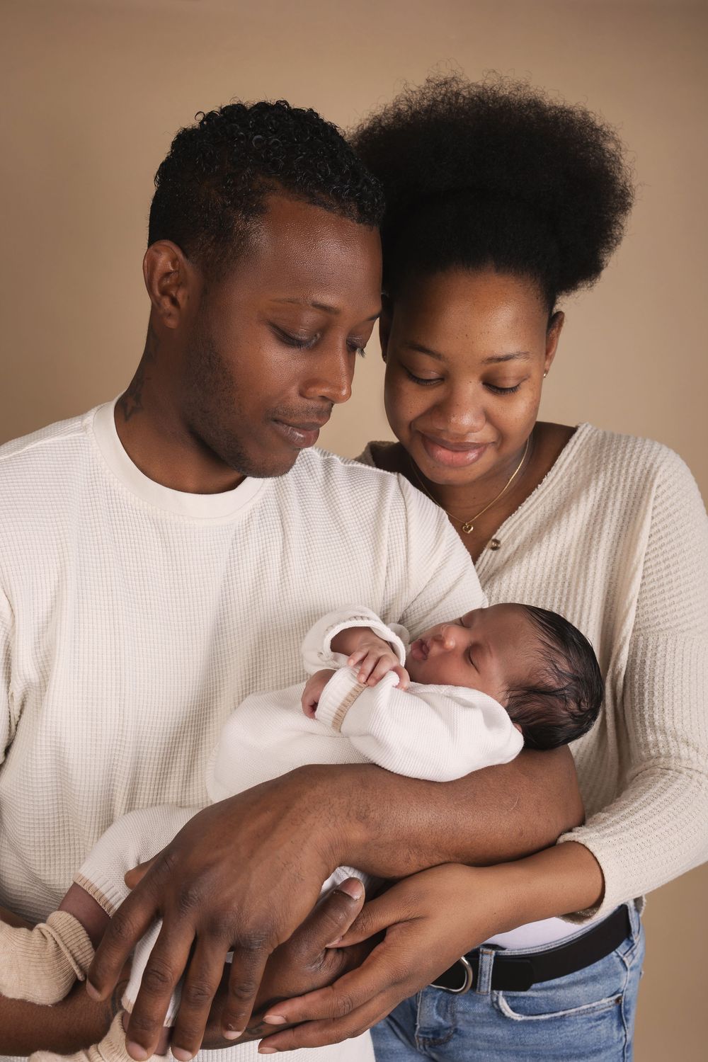 A tender family moment as parents in white sweaters lovingly gaze at their sleeping newborn baby swaddled in white.