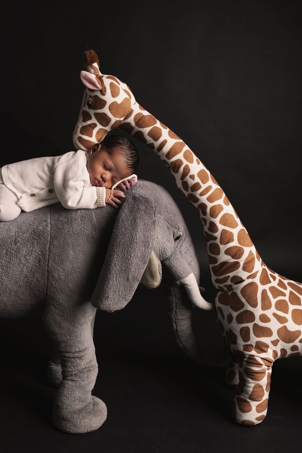 Sleeping baby nestled between large plush elephant and giraffe stuffed animals against dark background.