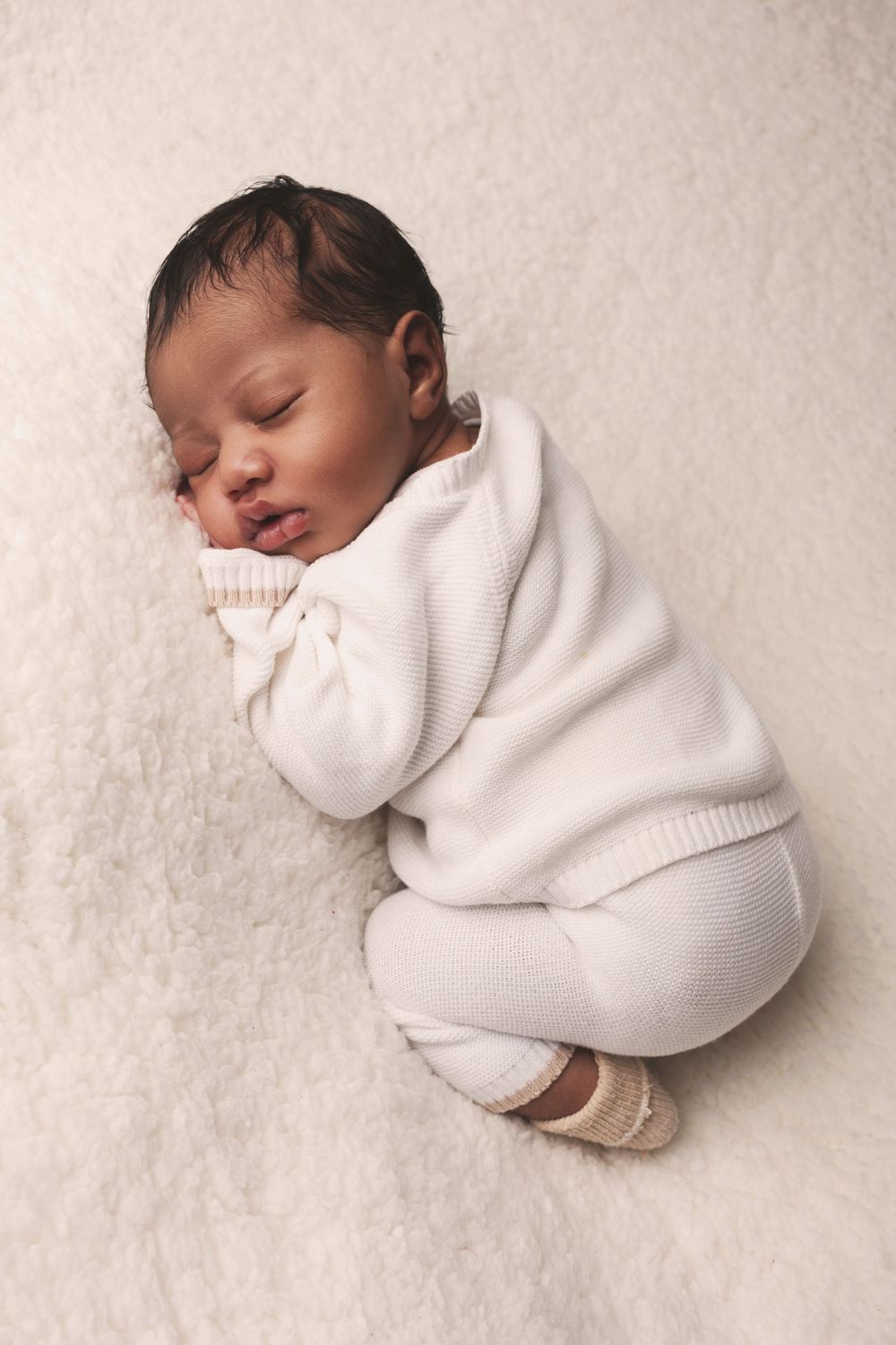 Sleeping newborn in cozy white knitted sweater and pants curled up on soft cream-colored blanket.