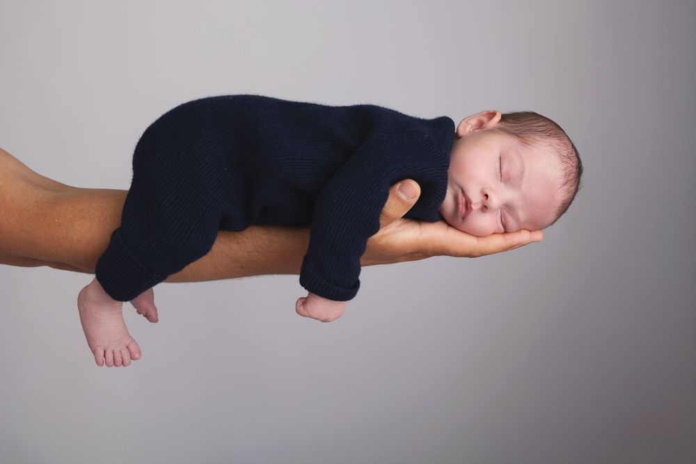 A sleeping newborn in a dark blue sleeper outfit rests peacefully on a supporting arm against a light gray background.