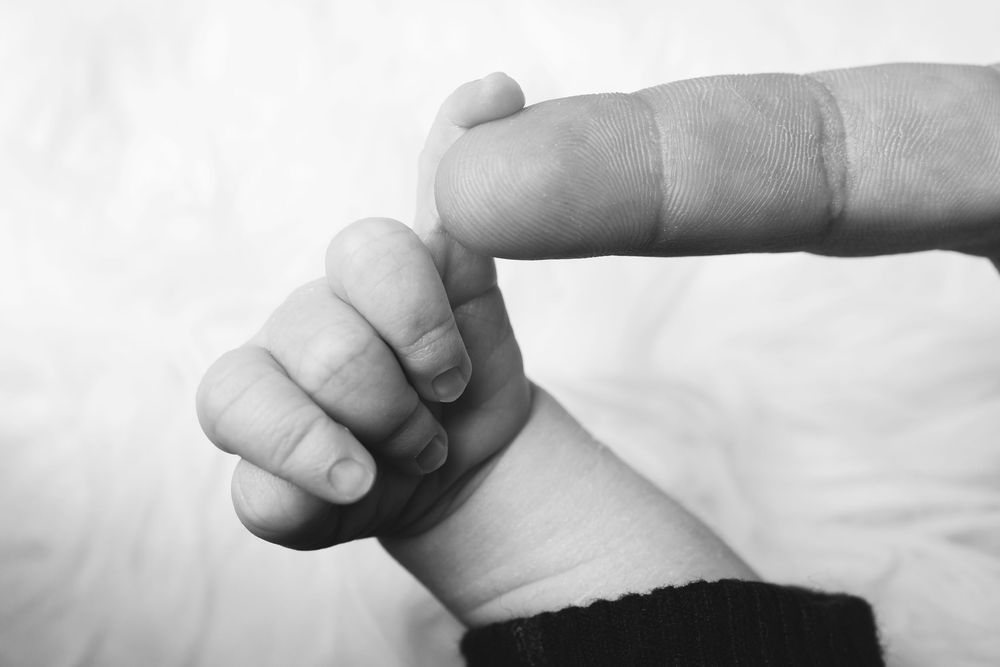 A black and white close-up photo shows a tiny baby's hand grasping an adult's finger against a light background.