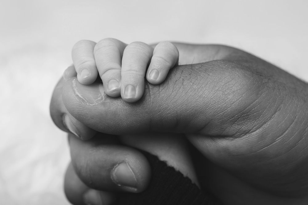 A small baby's hand grasps tightly onto an adult's finger in black and white.