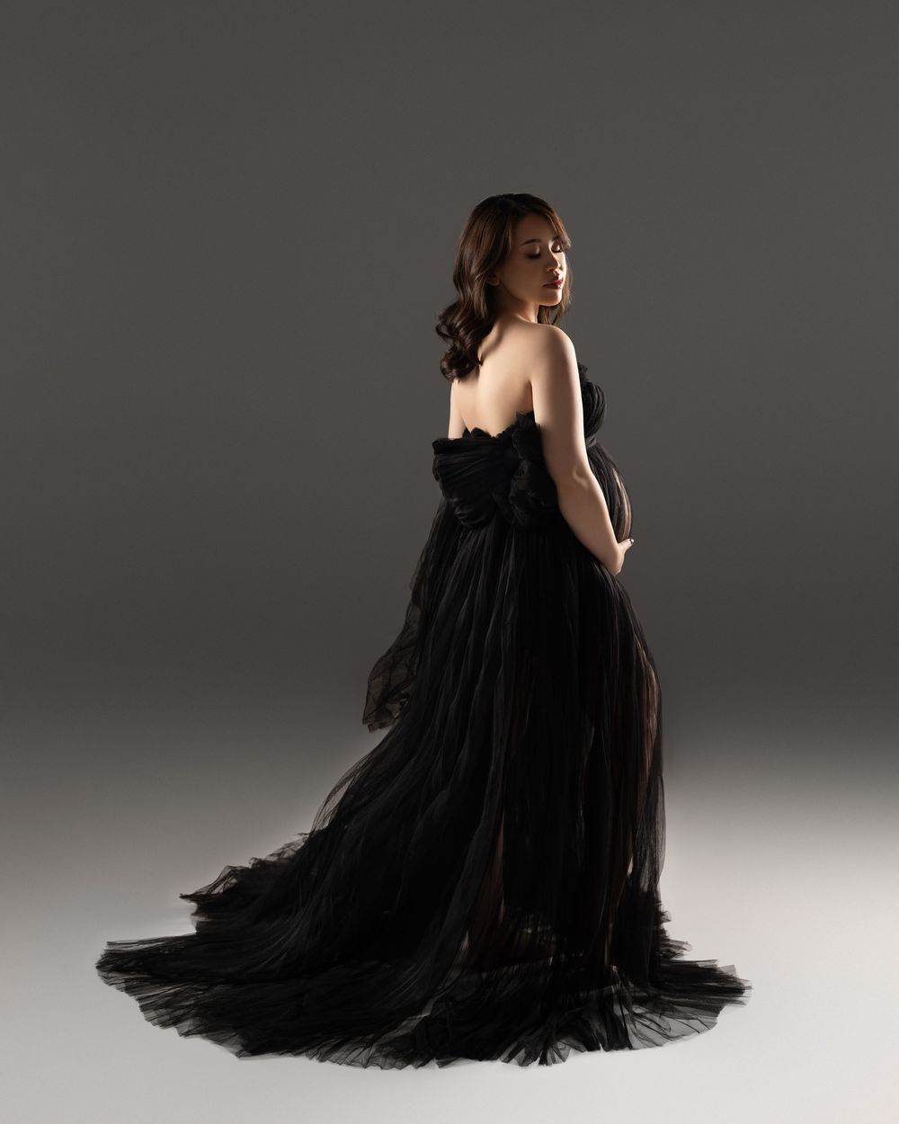 Elegant model in flowing black tulle gown poses sideways against dark grey studio backdrop with dramatic lighting.
