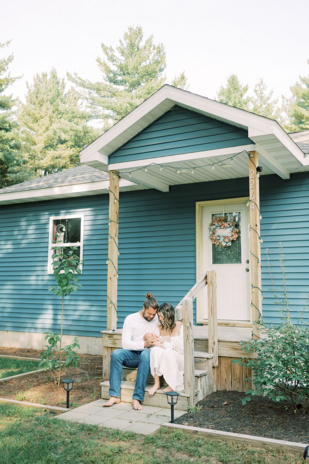 Couple sitting on porch steps of blue house with white trim and evergreen trees.