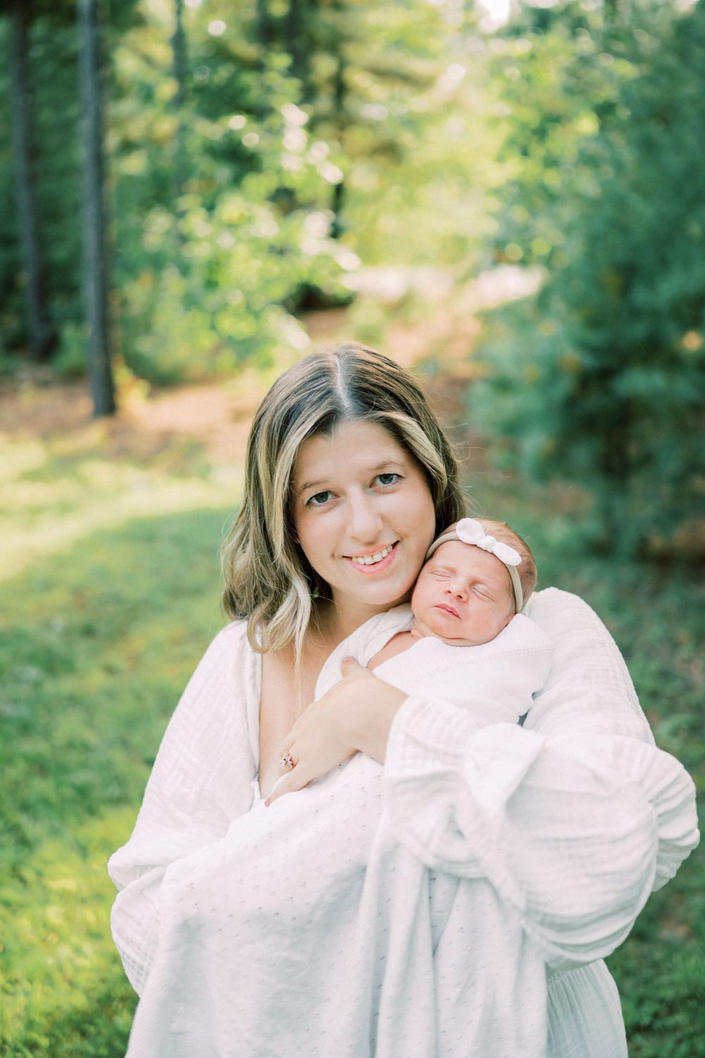 Outdoor portrait in sunlit forest with sleeping newborn in white.