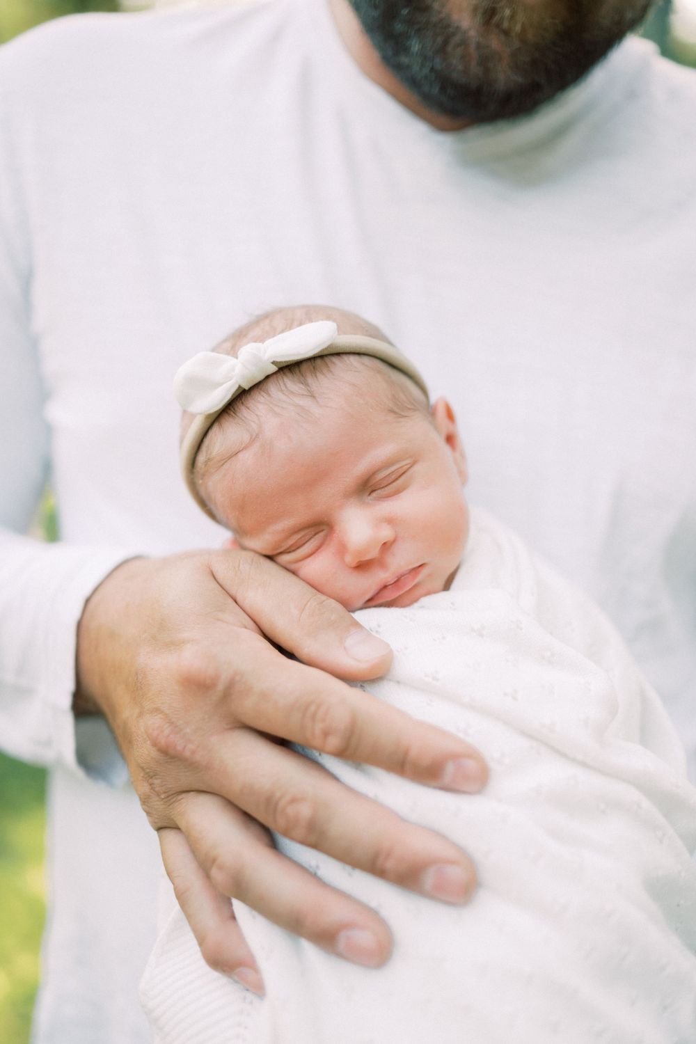 Sleeping newborn with white headband held gently against white shirt.