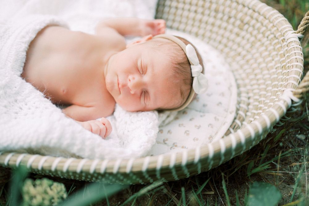 Peaceful sleeping newborn with headband resting in woven basket outdoors.