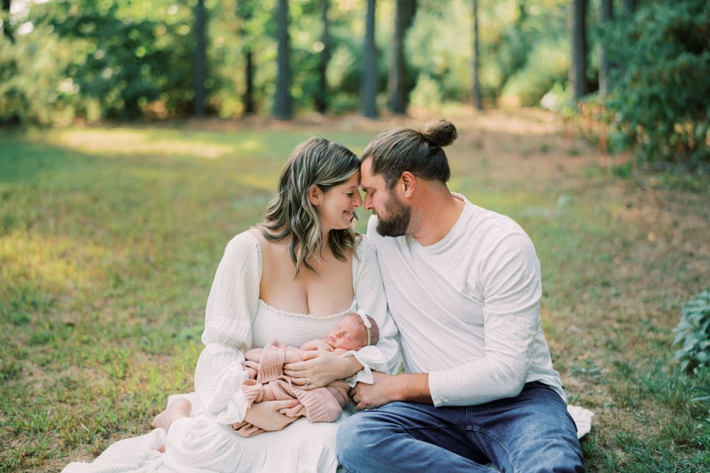 Couple sits on grass in wooded setting while holding their newborn in warm afternoon light.