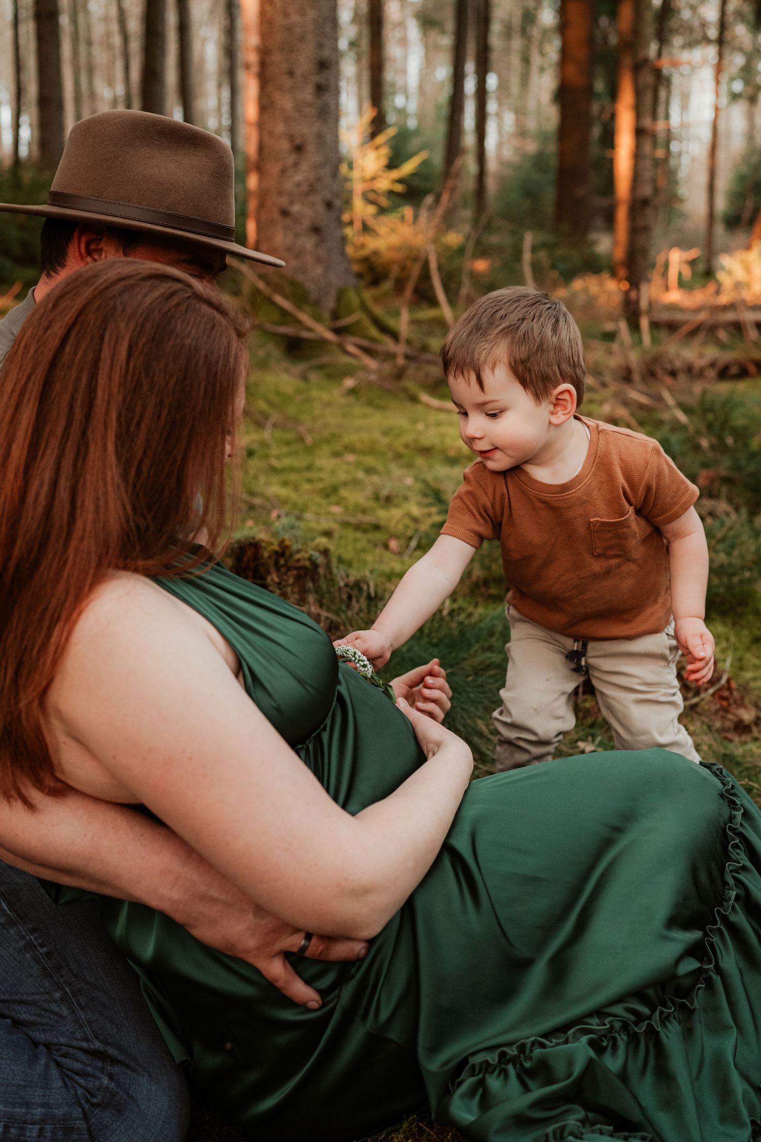 A child in a brown shirt reaches toward a woman wearing a green dress and brown hat as they sit together in a forest setting.