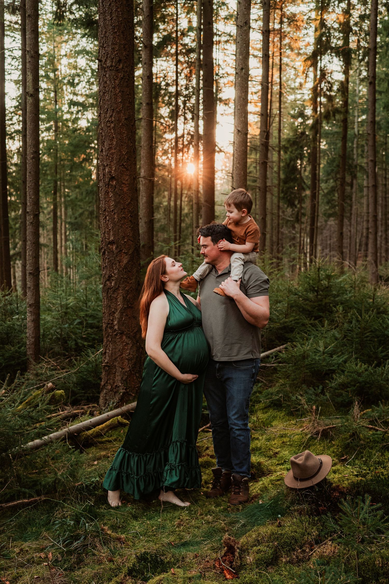 A couple shares a tender moment in a sunlit forest at sunset while their small child sits on shoulders.