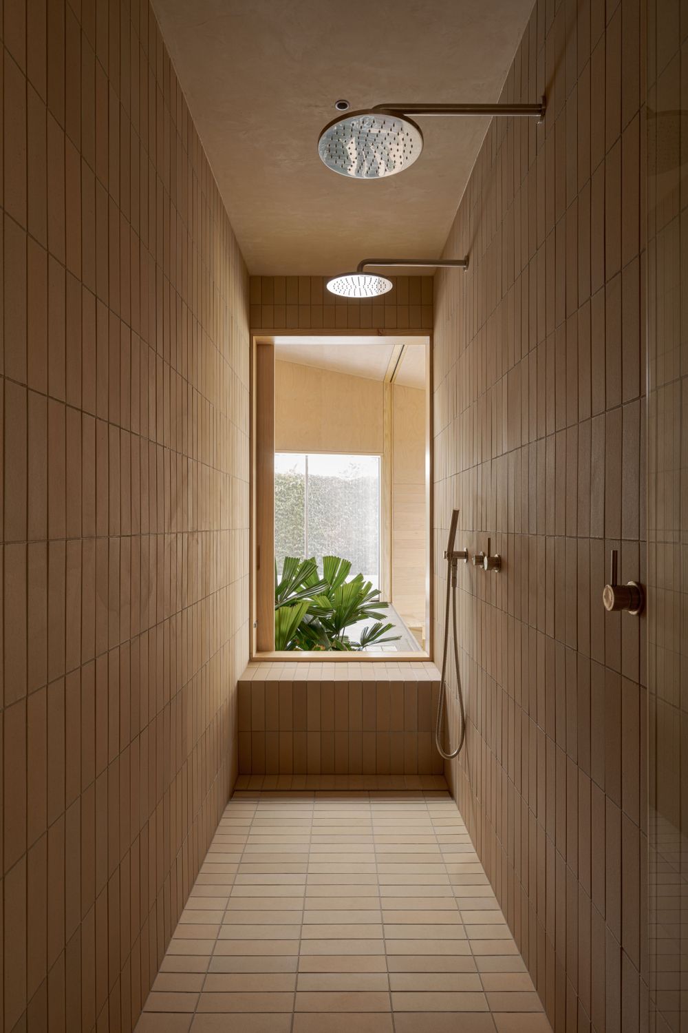 Modern bathroom hallway with beige tiles, rainfall shower head, and window overlooking tropical plants.