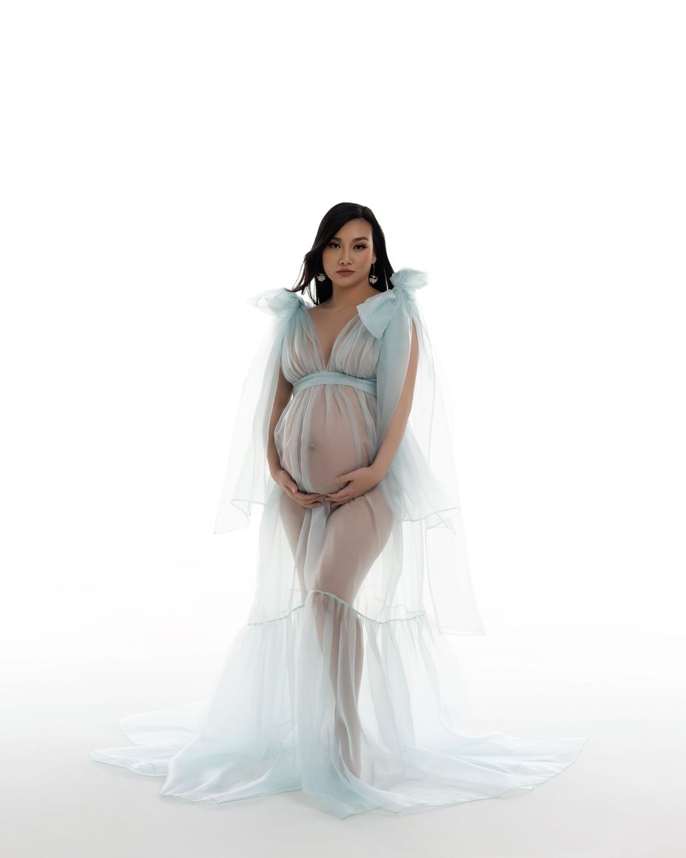 A pregnant woman poses in a flowing white maternity gown against a bright white studio backdrop.