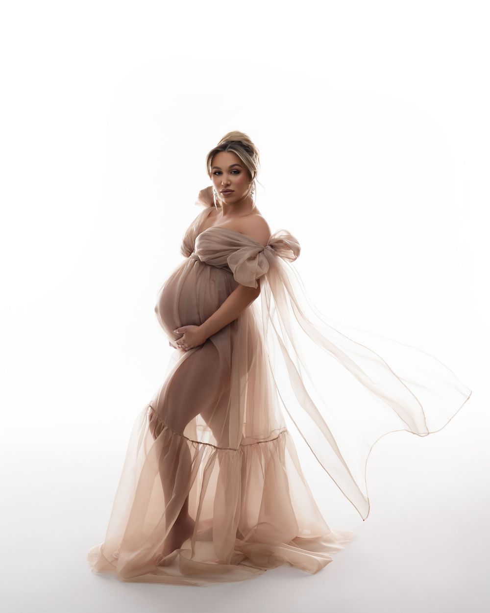 Elegant maternity portrait with flowing sheer blush gown against white background capturing a graceful maternal moment.