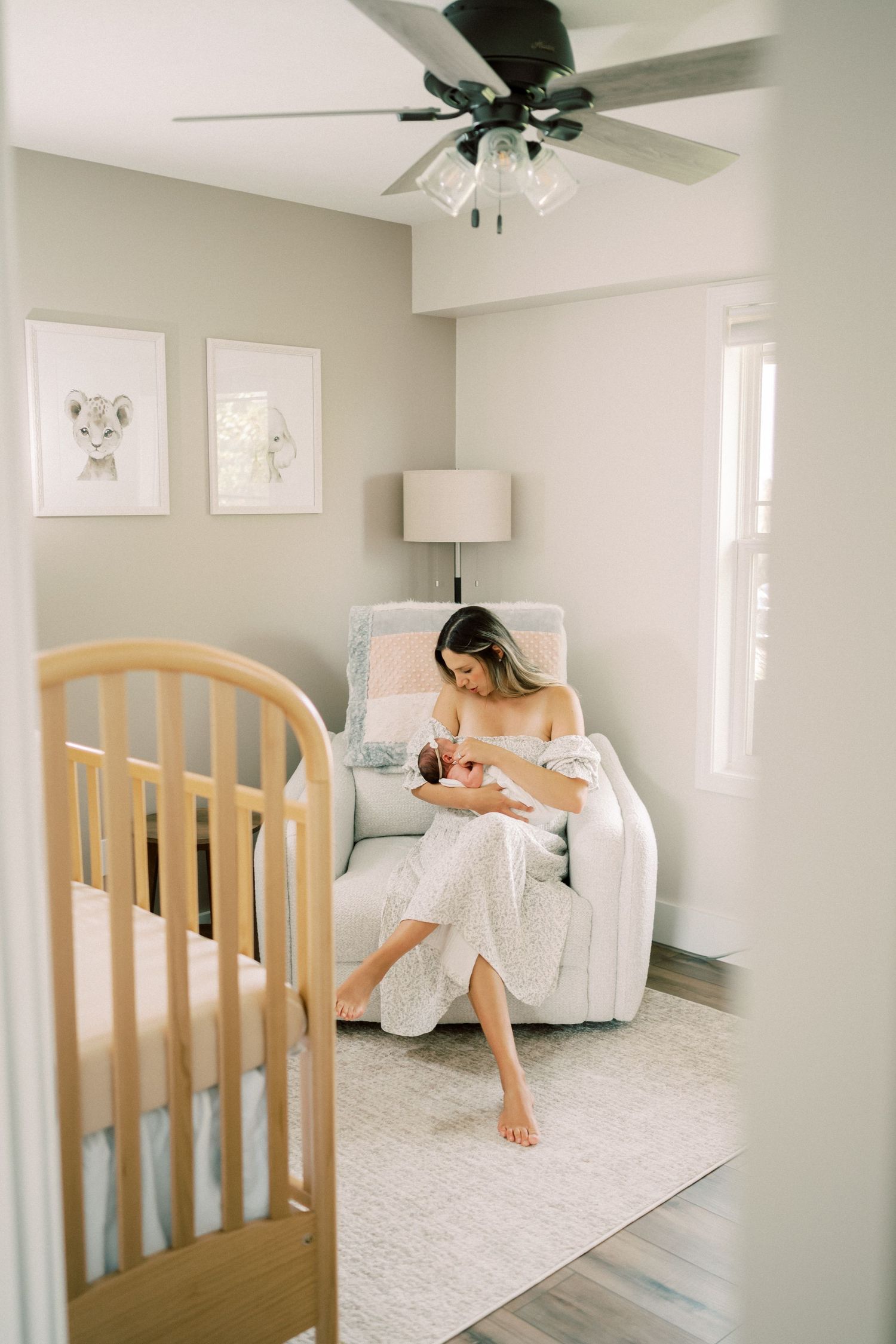 A peaceful nursery room with a wooden crib, glider chair, light gray walls, and ceiling fan with lamp fixtures.