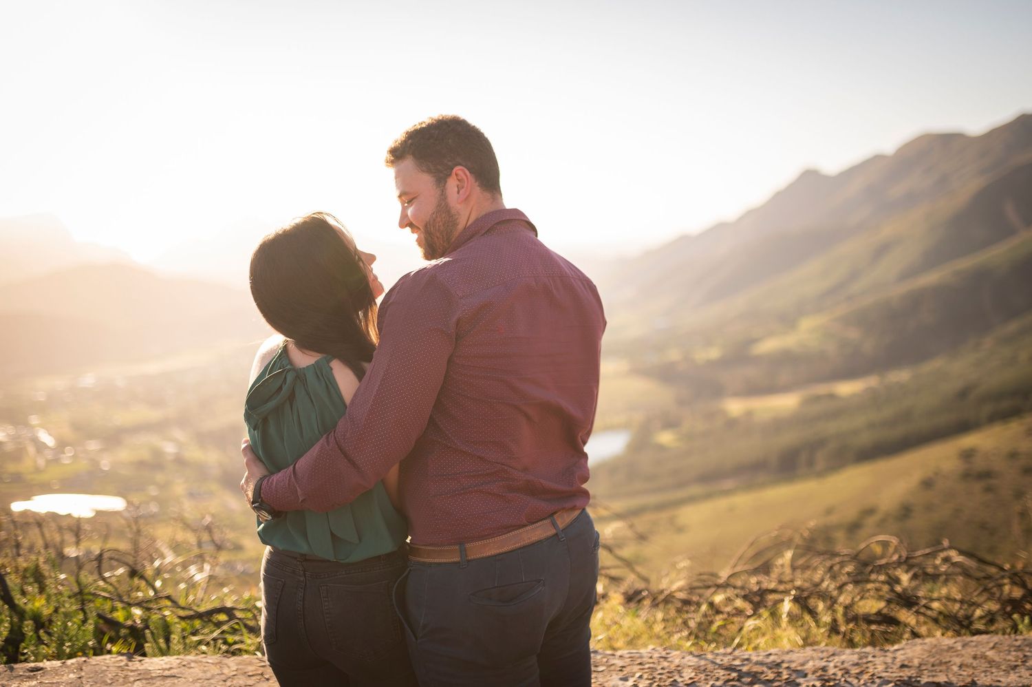 A couple embraces on a hilltop overlooking a scenic mountain valley landscape during golden hour sunset.