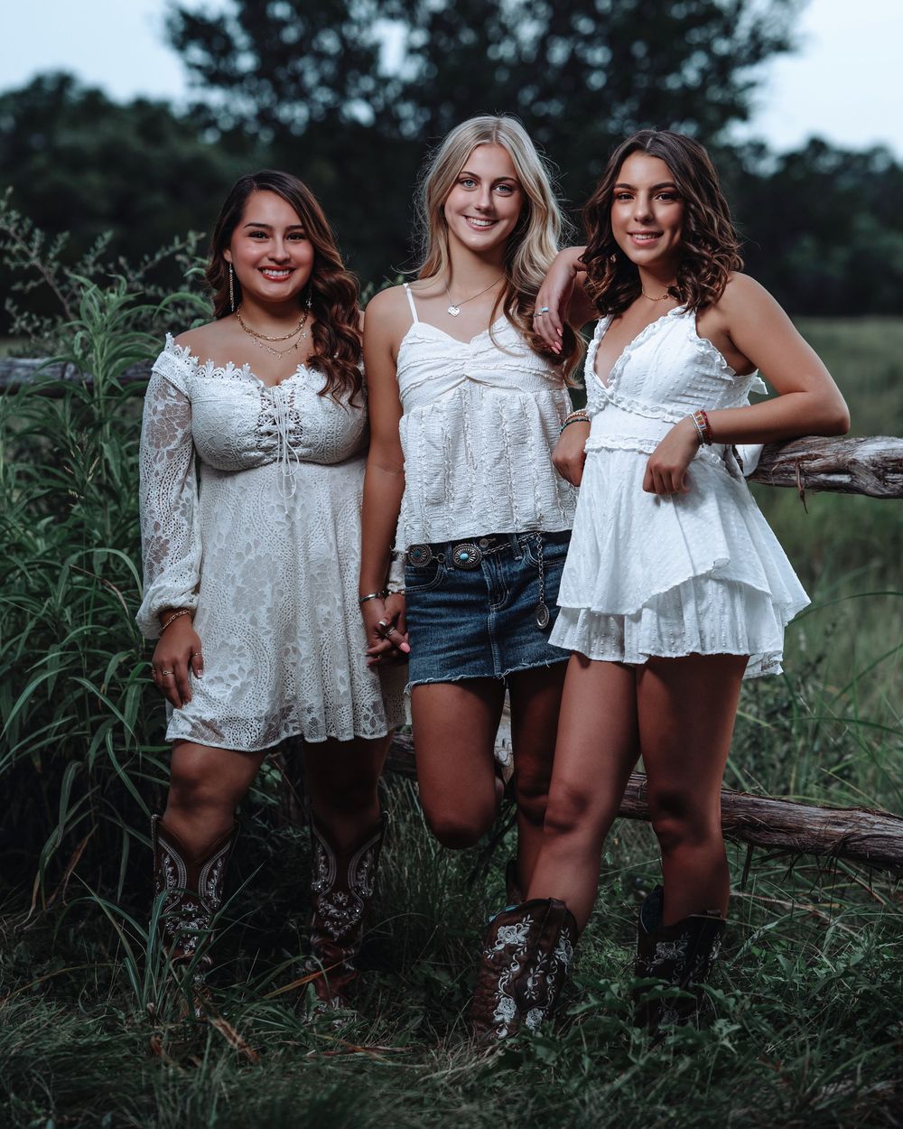 Three friends in white dresses and cowboy boots pose together in a rustic outdoor setting.