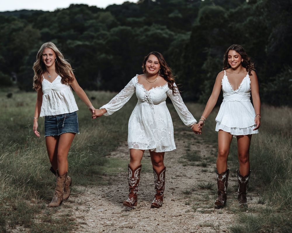Three friends in white tops and denim shorts hold hands while walking down a gravel path in a rustic outdoor setting.