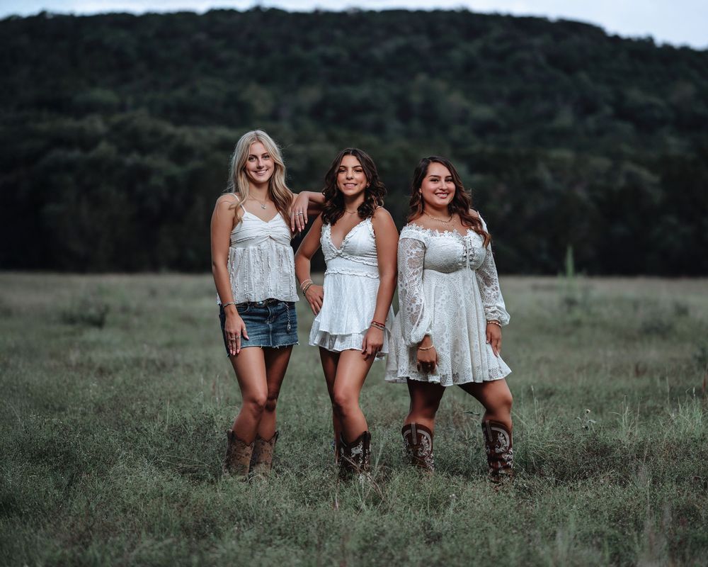 Three friends wearing white tops and cowboy boots pose together in a grassy field at sunset.