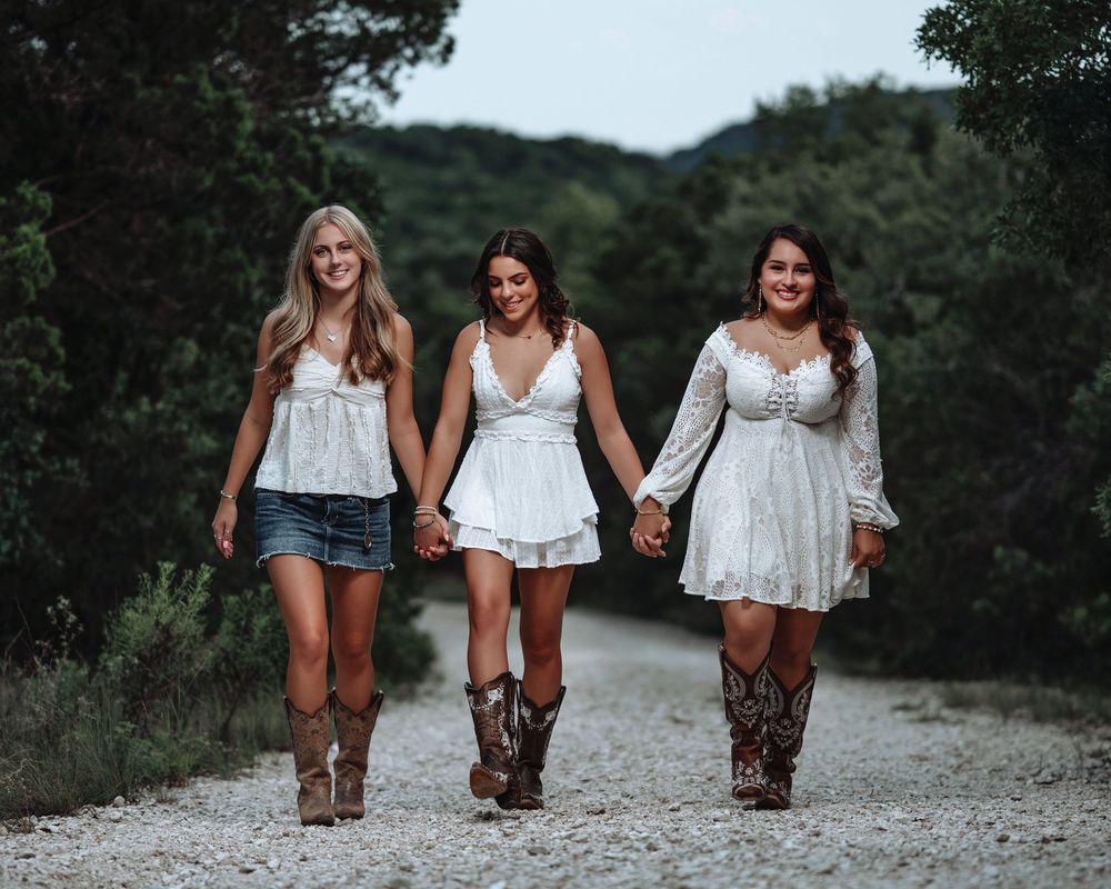 Three women in matching white outfits and cowboy boots walk along a country path surrounded by greenery.