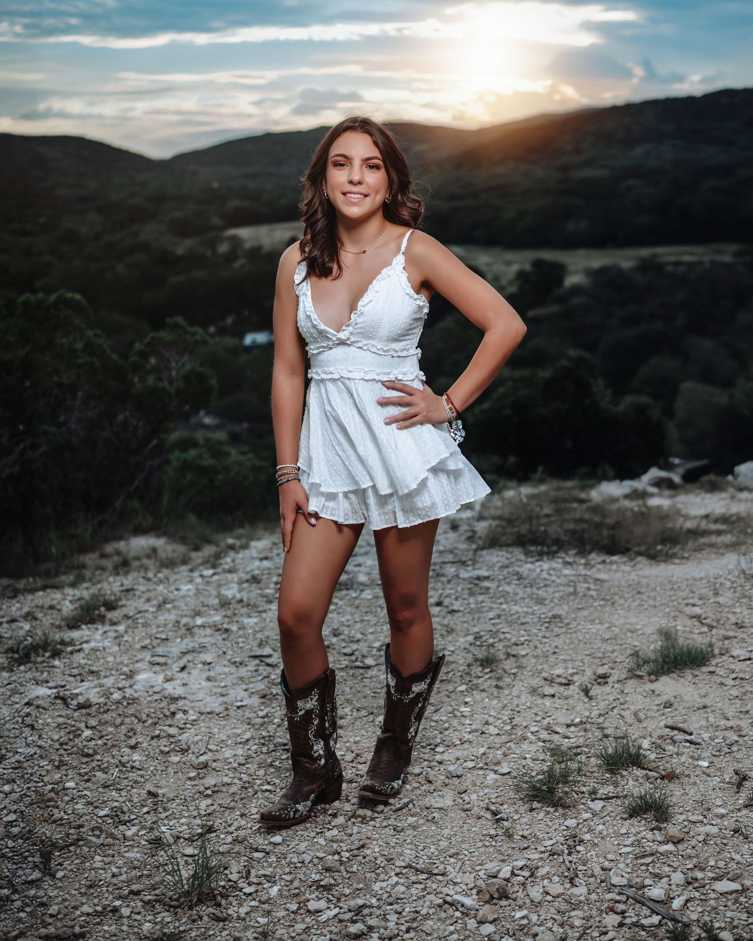 Someone wearing cowboy boots and denim shorts sits on a rocky cliff edge during sunset with mountains in the background.