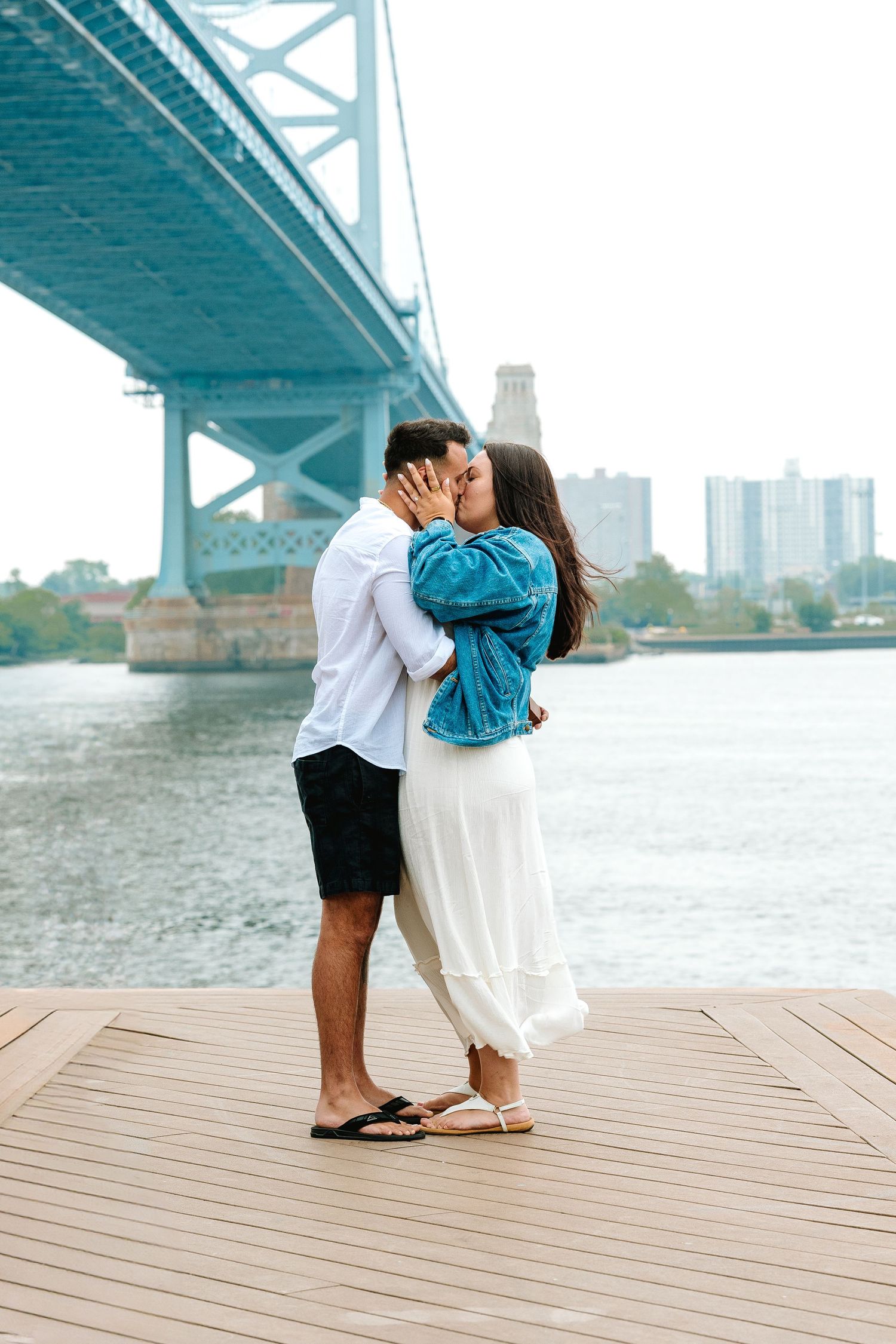 A romantic couple shares an intimate moment under a turquoise bridge near the water on a cloudy day.