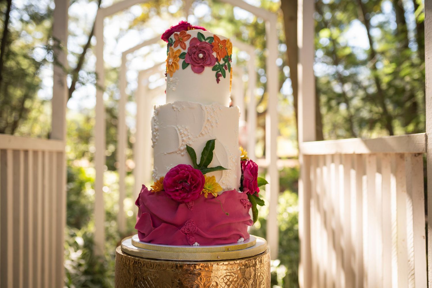 An elegant white wedding cake decorated with red flowers and gold leaf accents displayed in an outdoor garden setting.