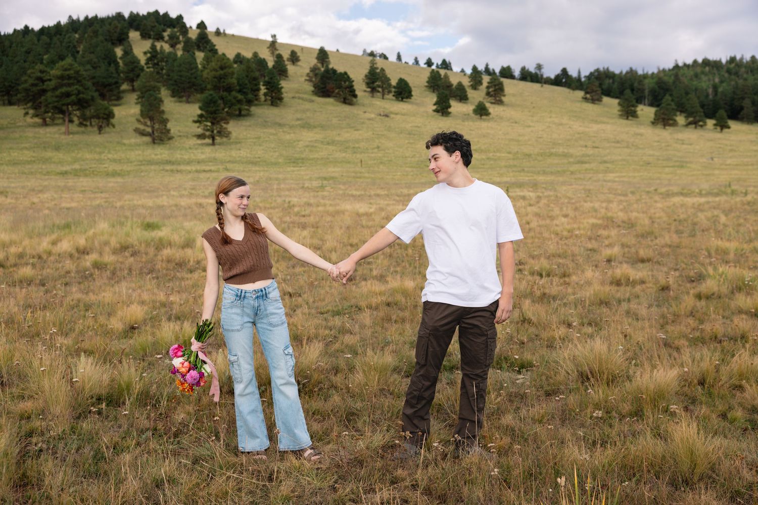 A couple is standing in a scenic mountain meadow with evergreen trees and cloudy skies in the background.