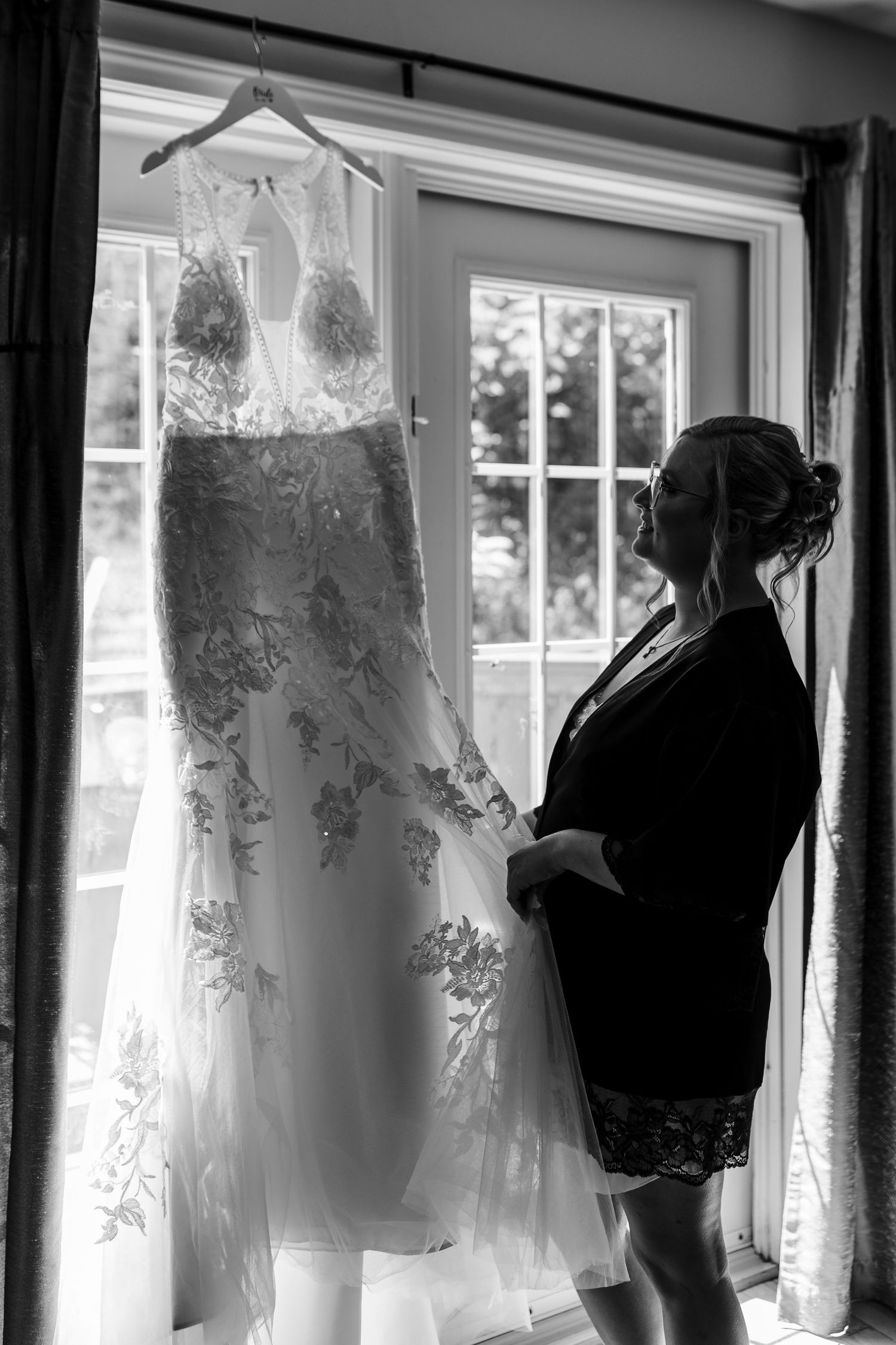 A delicate lace wedding dress hangs by a window as a silhouetted figure admires it in black and white.