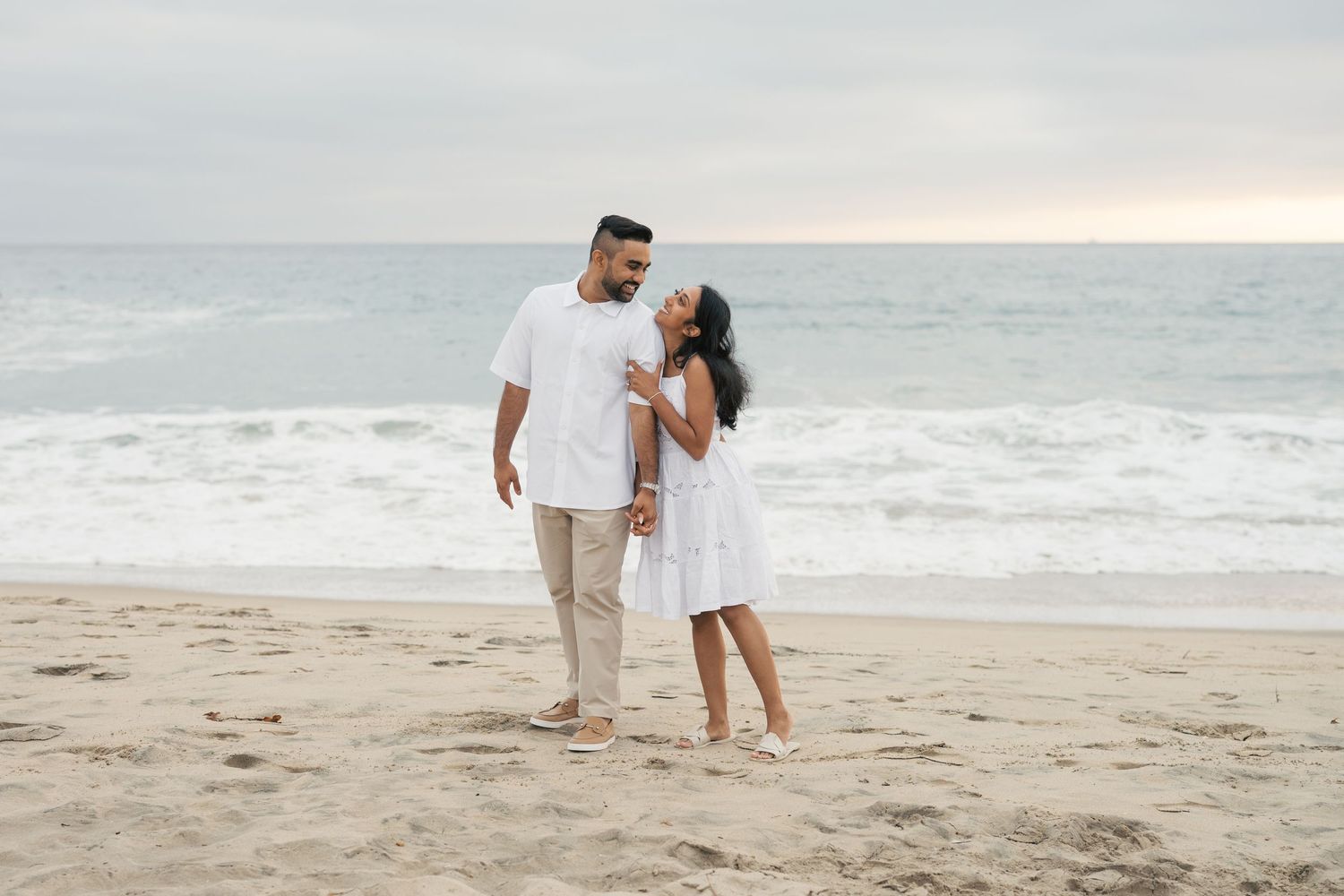 Romantic couple shares tender moments on sandy beach at sunset with ocean waves in background.