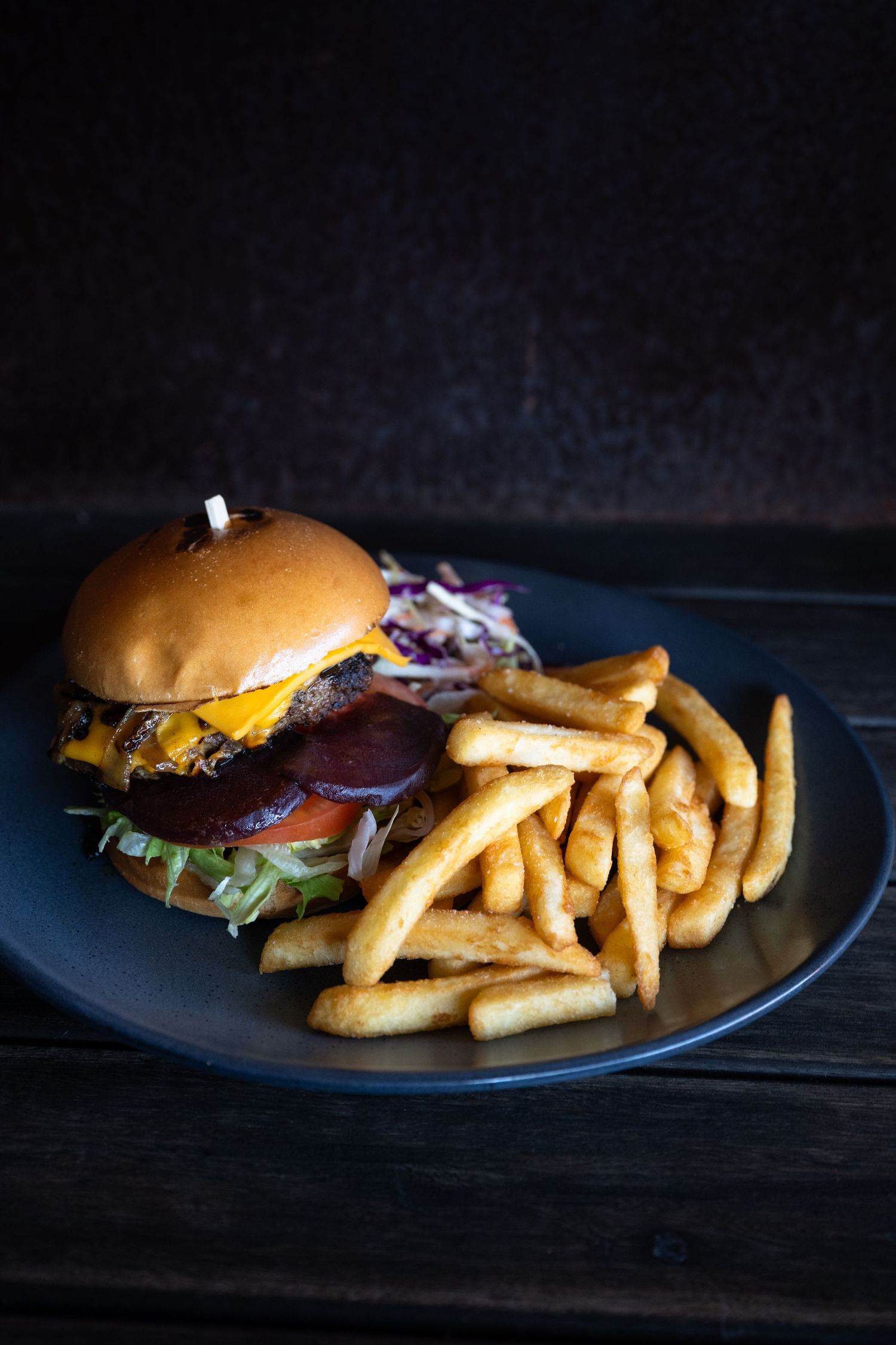 A juicy cheeseburger with toppings served on a black plate with crispy golden french fries on a dark wooden surface.
