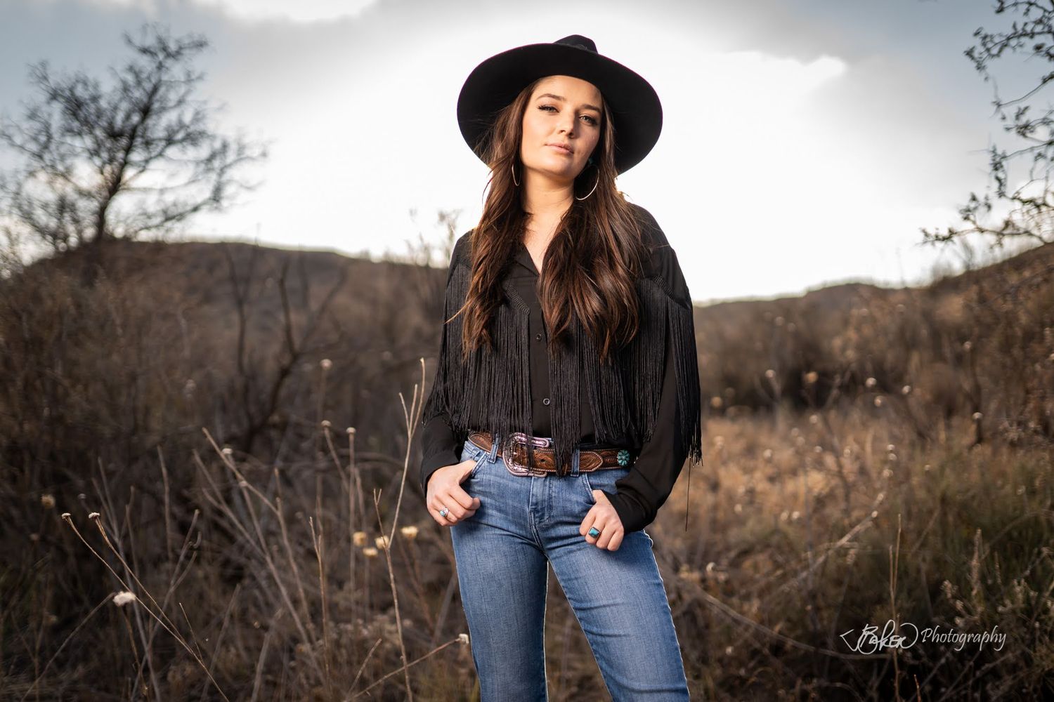 A western style portrait outdoors wearing a black shirt, jeans and wide brim hat against a mountain landscape.