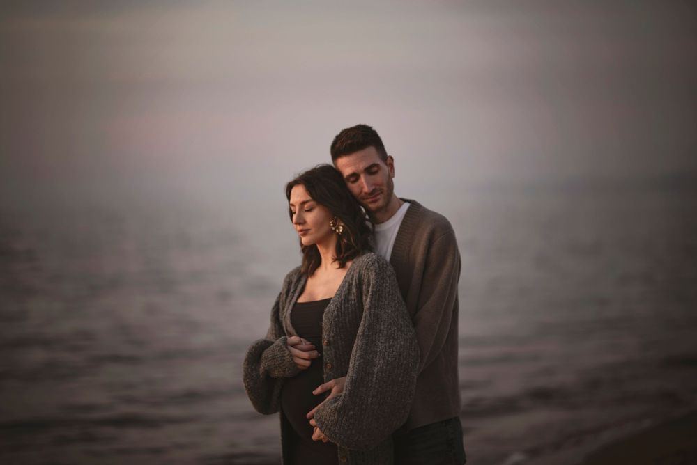 Romantiskt par omfamnas på en dimmig strand i solnedgången – en stämningsfull gravidfotografering i Skåne nära Hässleholm.
