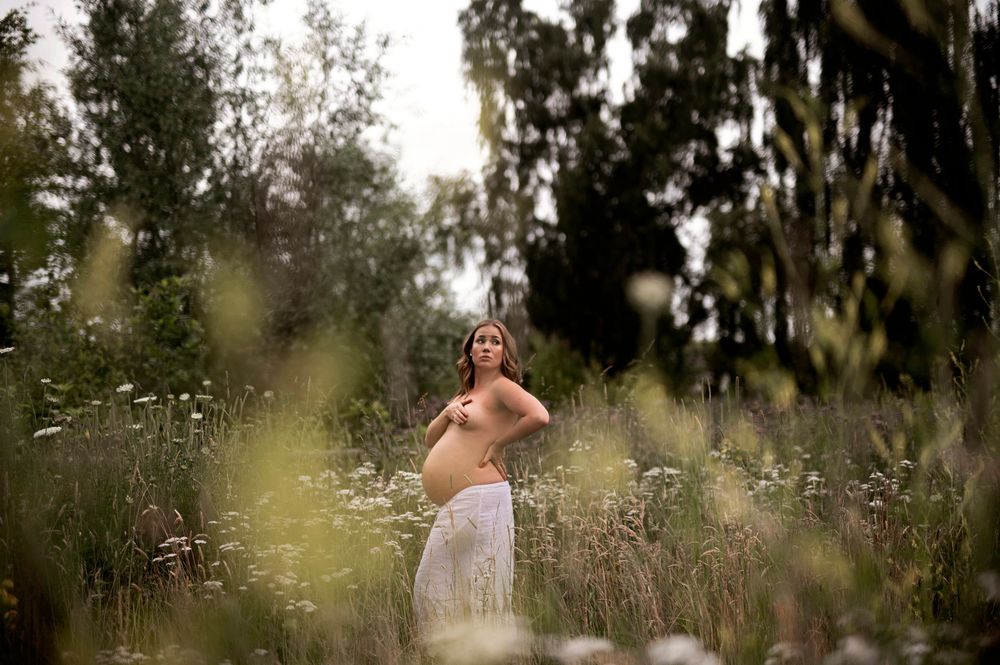 Gravid kvinna, vit klänning poserar i högt gräs och träd, i en drömmig utomhusmiljö – perfekt för gravidfotografering i Skåne