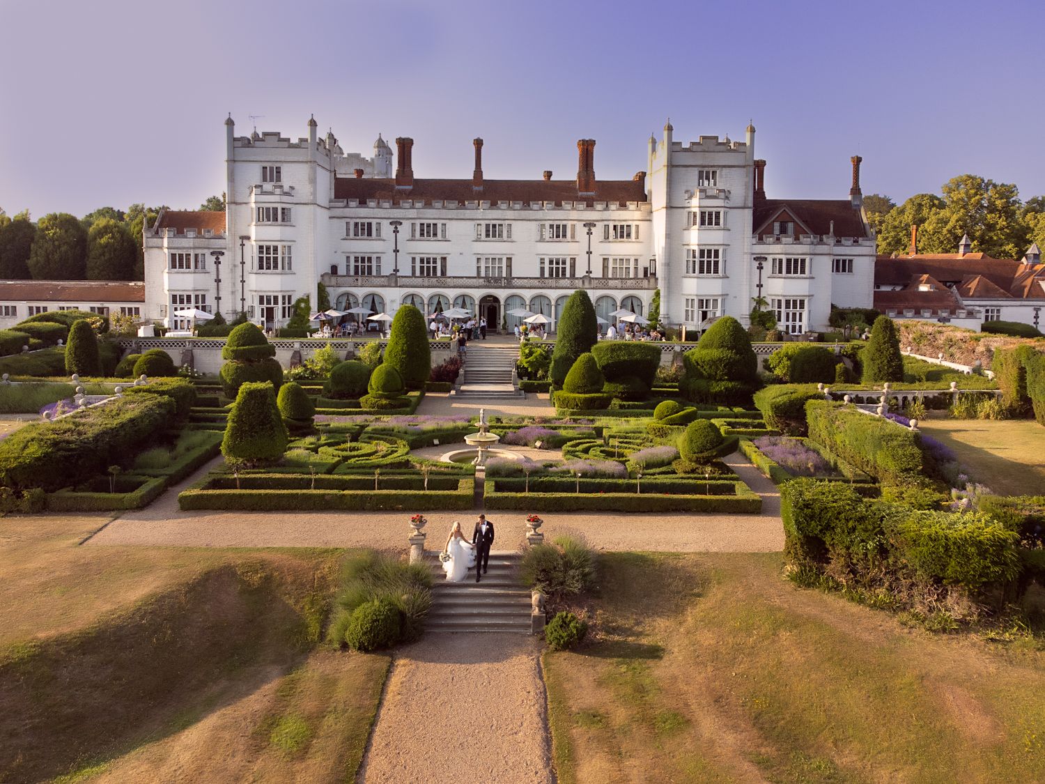 Historic white mansion overlooking manicured gardens and grand stone steps in a panoramic aerial view.