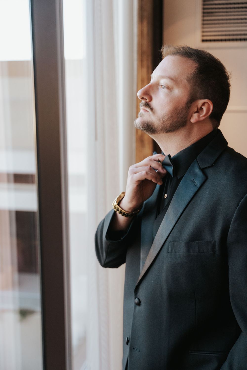 Professional in a dark suit adjusts his tie while contemplatively looking out a bright window.