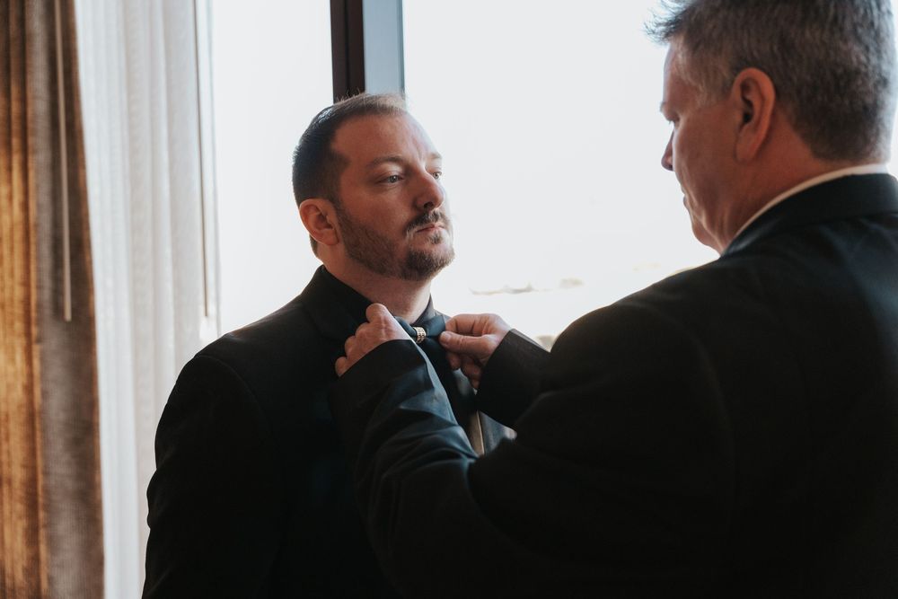 Two men in formal attire stand near a window while one adjusts the other's bow tie in an elegant setting.