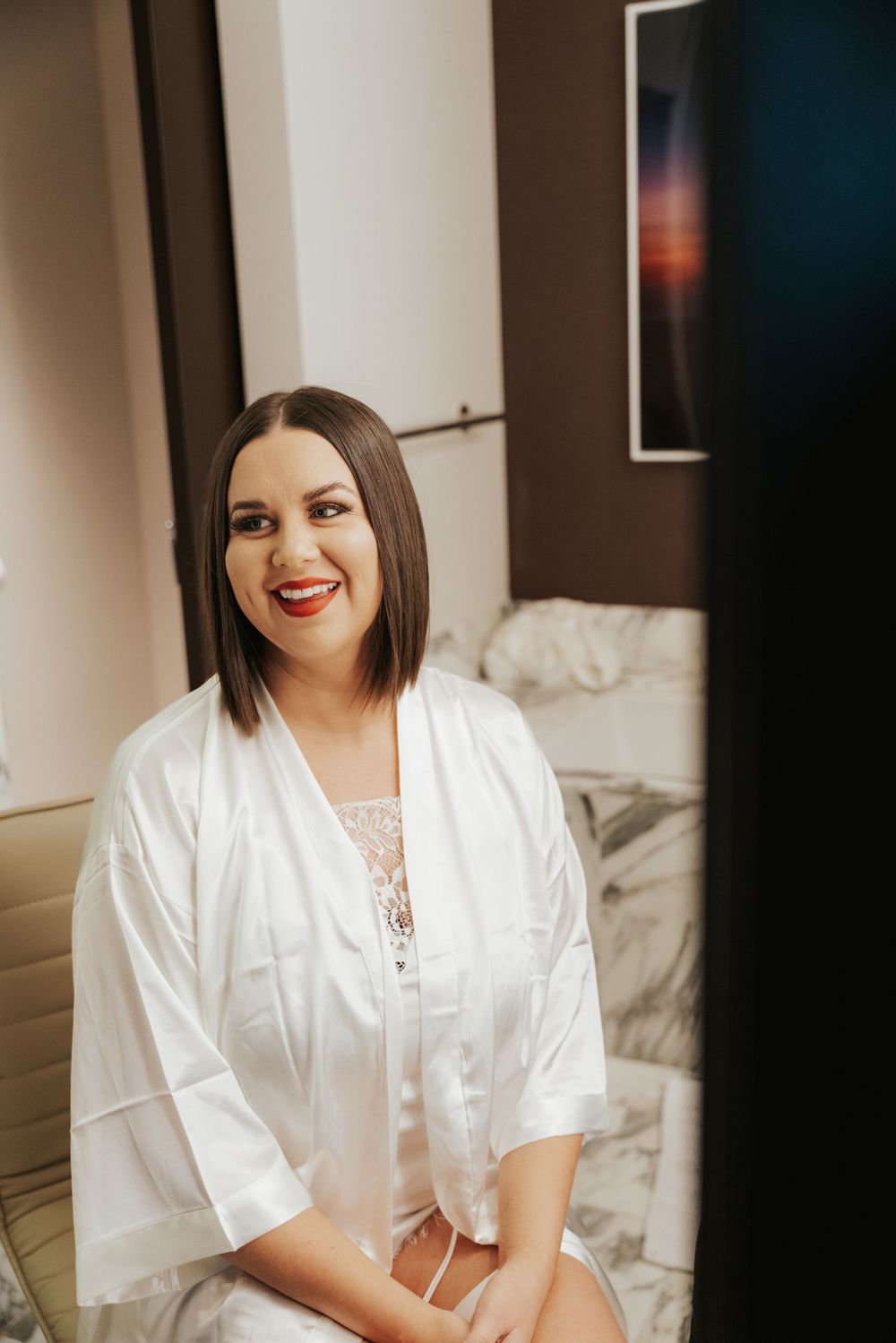 A joyful bride wearing a silky white robe sits on a bed during wedding preparation.