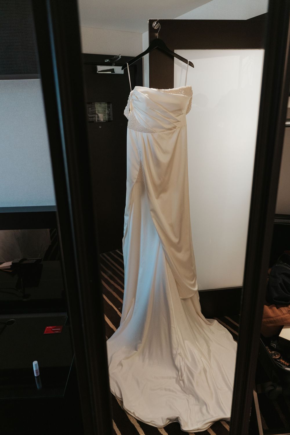 An elegant white wedding dress with draped fabric and flowing train hangs on a black garment rack against a light wall.