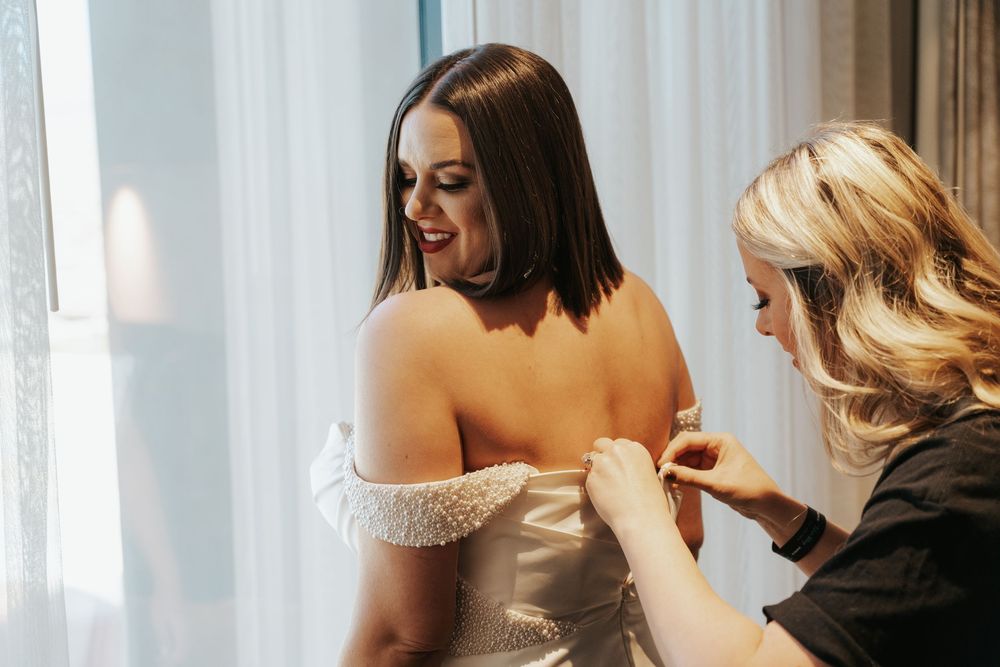 Someone helps adjust an off-shoulder wedding dress with delicate lace details near a window in natural light.